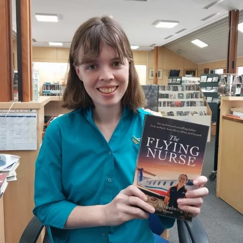 A library staff member stands in the library holding The Flying Nurse by Prudence Wheelwright