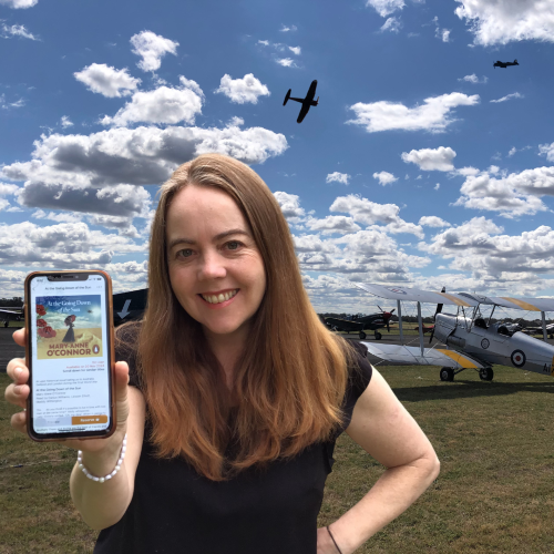 A library staff member displays At the Going Down of the Sun on an iPhone with an airshow and airfield in the background