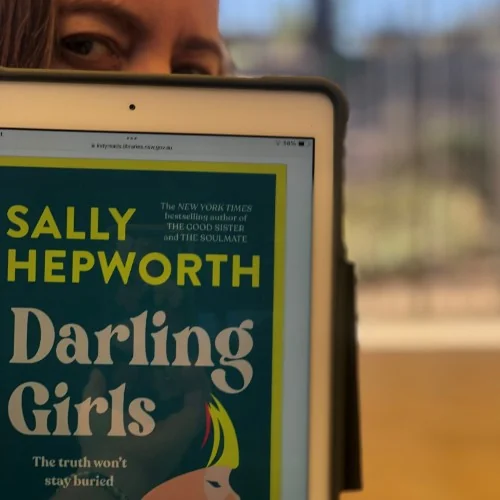 The eBook Darling Girls by Sally Hepworth is displayed on a tablet, with a library staff member peeking out behind.