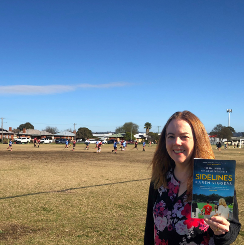 A library staff member stands on the sidelines of a junior soccer game, holding the book Sidelines by Karen Viggers