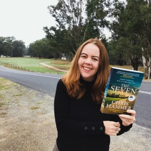 A Library staff member displays the book, The Seven by Chris Hammer with a country road in the background.