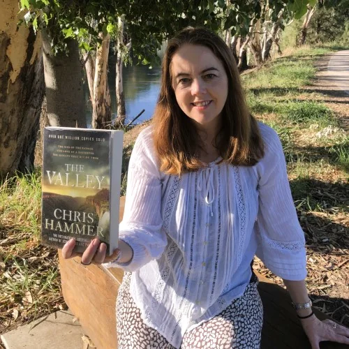 A library staff member holds up the book The Valley by Chris Hammer in an outdoor riverside setting.