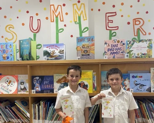 Two boys pose with their summer reading challenge bookmarks in front of the kids summer reading display.